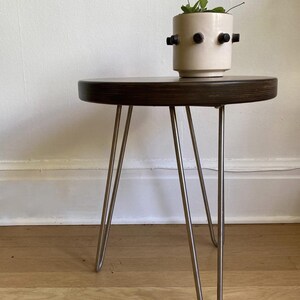 Puede incluir: Una mesa redonda de madera marr&oacute;n oscuro con patas de horquilla. Un macetero de cer&aacute;mica blanca con detalles negros se encuentra encima de la mesa.