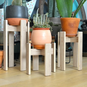 May include: Three wooden plant stands with different sized pots and plants. The stands are made of light wood and have a simple design. The pots are all different colors and styles.