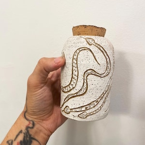 May include: A speckled white ceramic jar with a cork stopper. The jar is decorated with etched snake designs in a brown color. The jar is held by a person.