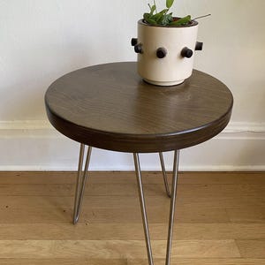 Puede incluir: Una mesa auxiliar redonda de madera con un tinte marr&oacute;n oscuro. La mesa tiene tres patas de horquilla de metal. Un macetero de cer&aacute;mica blanca con detalles negros se encuentra sobre la mesa.