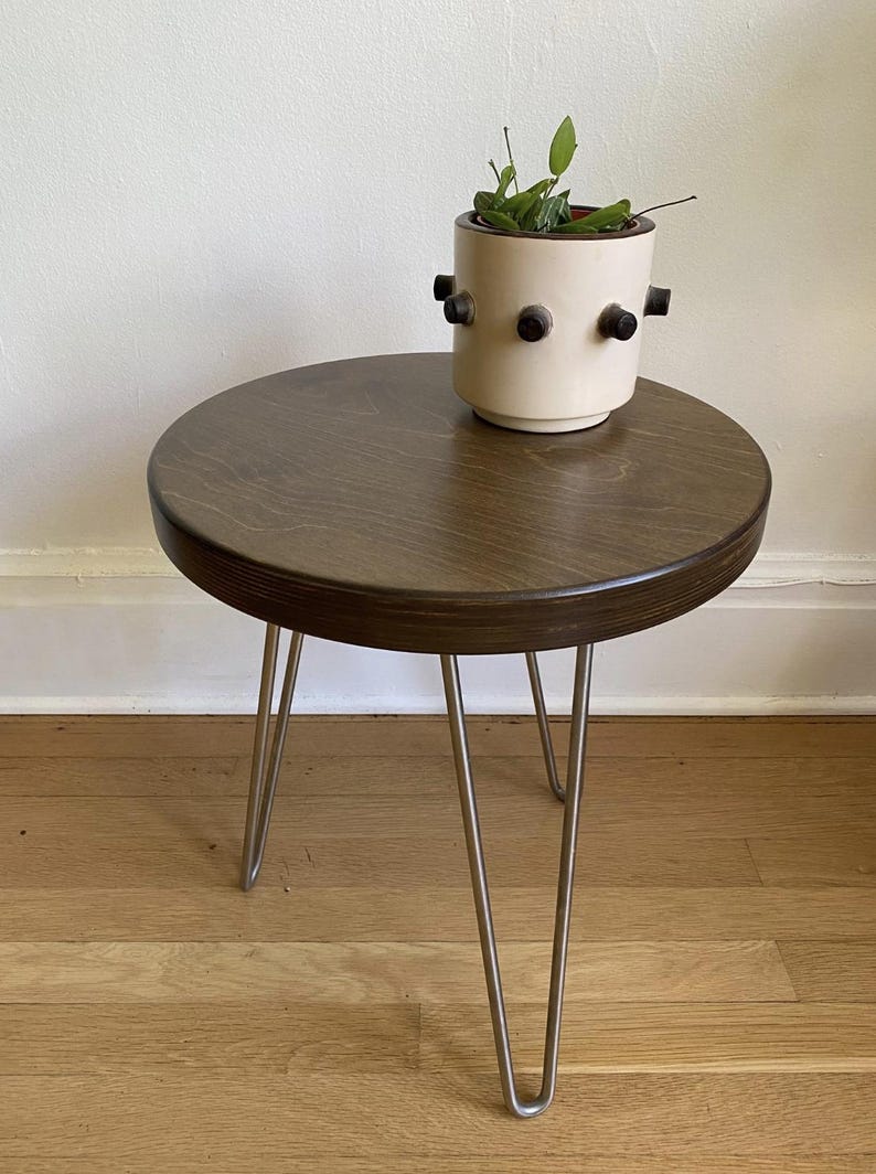 Puede incluir: Una mesa auxiliar redonda de madera con un tinte marr&oacute;n oscuro. La mesa tiene tres patas de horquilla de metal. Un macetero de cer&aacute;mica blanca con detalles negros se encuentra sobre la mesa.