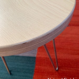 Puede incluir: Una mesa redonda de madera con un acabado natural y tres patas finas de metal. La mesa est&aacute; sobre una alfombra colorida con secciones rojas, amarillas, verdes y azules. El texto "Natural Finish" es visible en la esquina inferior derecha de la imagen.