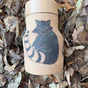 May include: A tan ceramic container with a wooden lid. The container features a black illustration of a raccoon with a striped tail. The container is cylindrical and sits on a bed of dried leaves.