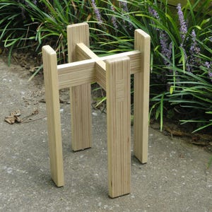 May include: A wooden plant stand made of plywood with a natural finish. The stand has four legs and a square top.