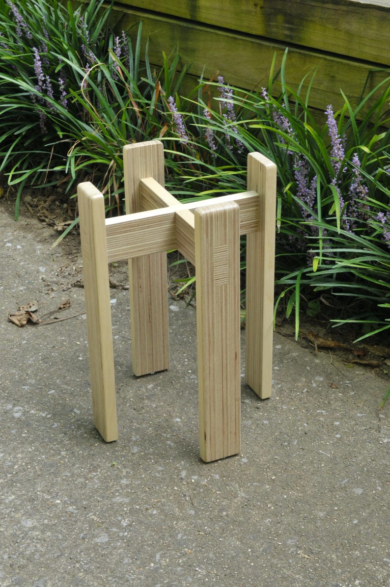 May include: A wooden plant stand made of plywood with a natural finish. The stand has four legs and a square top.