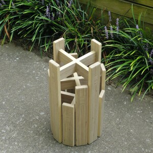 May include: A wooden plant stand made of plywood with a criss-cross design. The stand is made of multiple pieces of wood that are stacked on top of each other.