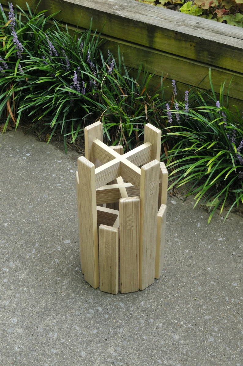 May include: A wooden plant stand made of plywood with a criss-cross design. The stand is made of multiple pieces of wood that are stacked on top of each other.