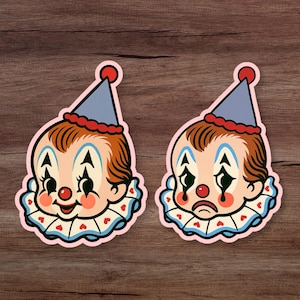 Puede incluir: Dos pegatinas con caras de payasos vintage. Uno sonríe, el otro está triste. Ambos tienen sombreros azules, adornos rojos y cuellos con volantes con detalles de corazón. Las pegatinas tienen un borde rosa y están sobre madera.