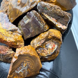 Puede incluir: Una colección de piedras de ópalo en bruto. Los ópalos exhiben una variedad de colores, incluyendo marrón, dorado y toques de azul y verde. Las piedras tienen patrones y texturas únicos, mostrando su belleza natural.