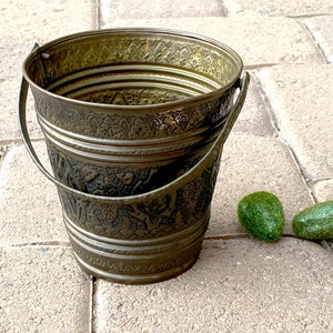 May include: A vintage-style metal bucket with a handle, featuring intricate embossed floral patterns. The bucket has a tarnished brass finish and is accompanied by two small, green, moss-covered decorative stones. The bucket is sitting on a stone surface.
