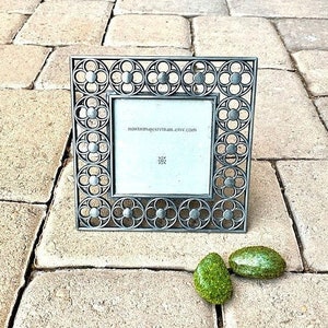 May include: A square, ornate metal picture frame with a floral design. The frame is silver-toned and has a small white insert. The text "nonimajestivam.etsy.com" is visible on the insert. Two small, green, oval-shaped decorative stones are next to the frame.