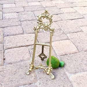 May include: A gold-colored, ornate easel with a decorative top and base. The easel is made of metal and has a detailed, vintage design. A small, green decorative ball is next to the easel. The easel is standing on a brick surface.