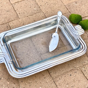 May include: A rectangular glass baking dish with a silver metal frame and handles. A silver serving utensil rests inside the dish. Two green decorative moss balls are on the side. The dish is on a brick surface.