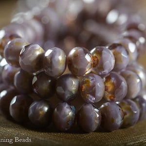 Puede incluir: Una hebra de cuentas de vidrio púrpura iridiscente con un acabado facetado. Las cuentas están dispuestas en un patrón circular, creando un efecto de capas.