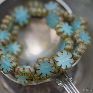 May include: A close-up of a strand of turquoise blue and green glass beads in the shape of flowers. The beads are arranged in a circle and are sitting in a silver bowl.