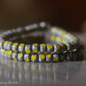 May include: Close-up of a bracelet made of small, round beads. The beads are primarily gray with yellow accents. The bracelet is doubled, with two strands visible. The background is blurred, with a dark, neutral tone.