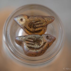 May include: Two gold-coloured ceramic bird beads with a textured surface. The beads are in a clear glass container.