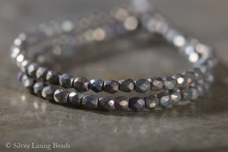 Puede incluir: Primer plano de una hebra de cuentas de vidrio facetadas de color gris oscuro. Las cuentas est&aacute;n dispuestas en un patr&oacute;n circular, creando un brazalete o un collar.