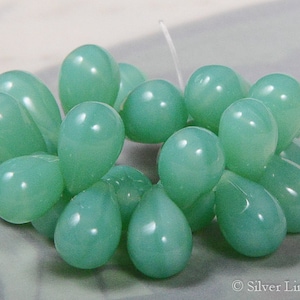 Puede incluir: Un grupo de cuentas de color verde claro en forma de lágrima, dispuestas en un patrón circular. Las cuentas están hechas de vidrio y tienen un acabado liso y pulido.