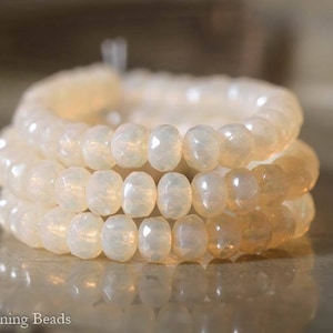 May include: A close-up of a stack of white and cream colored glass beads. The beads are faceted and have a slightly iridescent finish.