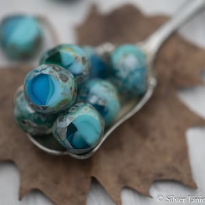 May include: Close-up of a collection of turquoise and blue glass beads with faceted surfaces, displayed in a silver spoon. The beads have a marbled appearance and are resting on a brown leaf.