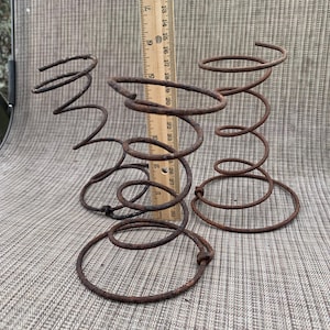 May include: Three rusty metal springs of varying sizes, arranged on a textured surface. The springs are a dark brown color and appear to be vintage or reclaimed. A wooden ruler is placed next to the springs for scale.
