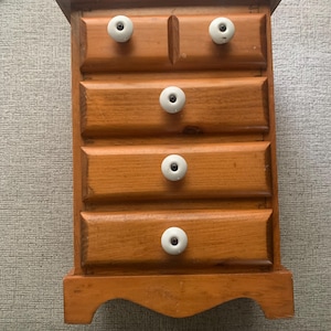May include: A small wooden chest of drawers with five drawers. The chest is stained a warm brown color and features white ceramic knobs. The bottom of the chest has a decorative scalloped edge.
