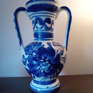 May include: A white ceramic vase with blue floral designs and two handles. The vase has a rounded body and a flared base.