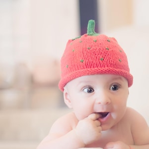 Peut inclure: Un b&eacute;b&eacute; portant un bonnet en tricot rouge en forme de fraise, regardant vers la droite, avec un doigt dans la bouche.