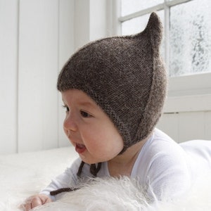 Könnte beinhalten: Ein Baby trägt eine braune Strickmütze mit einem spitzen Ohrenklappen. Die Mütze ist aus weichem, strukturiertem Garn gefertigt. Das Baby liegt auf einer weißen, flauschigen Oberfläche.