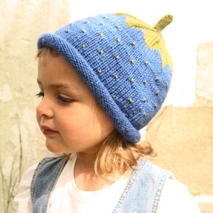 Peut inclure: Un enfant portant un bonnet tricot&eacute; bleu et vert en forme de fraise. Le bonnet a une tige verte et des graines jaunes.