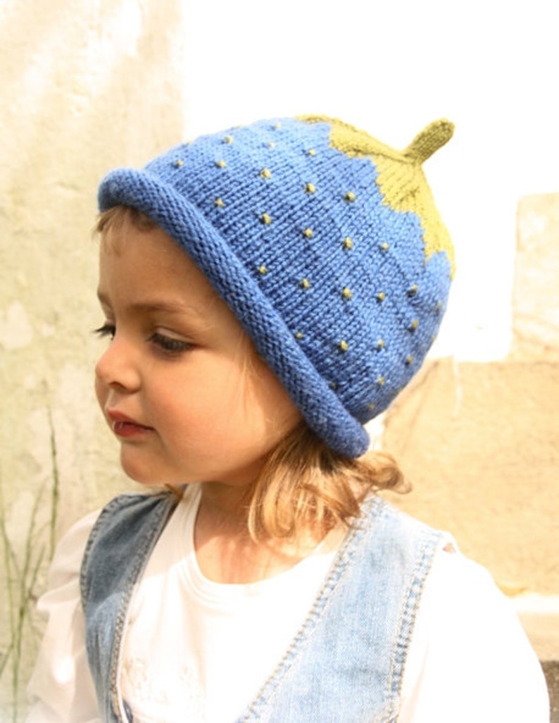 Peut inclure: Un enfant portant un bonnet tricot&eacute; bleu et vert en forme de fraise. Le bonnet a une tige verte et des graines jaunes.