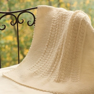May include: A cream-colored crocheted baby blanket with a delicate, lacy pattern. The blanket is draped over a black metal chair with a blurred background of fall foliage.