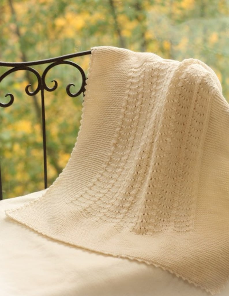 May include: A cream-colored crocheted baby blanket with a delicate, lacy pattern. The blanket is draped over a black metal chair with a blurred background of fall foliage.