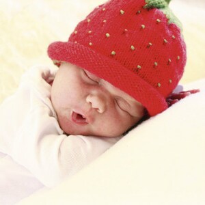 Peut inclure: Un b&eacute;b&eacute; portant un bonnet en tricot rouge en forme de fraise avec des feuilles vertes dort sur une couverture blanche.