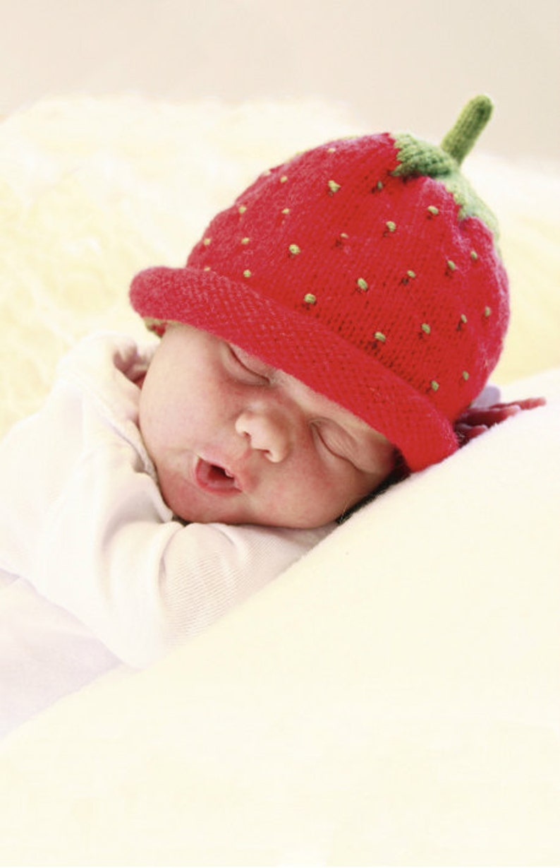 Peut inclure: Un b&eacute;b&eacute; portant un bonnet en tricot rouge en forme de fraise avec des feuilles vertes dort sur une couverture blanche.