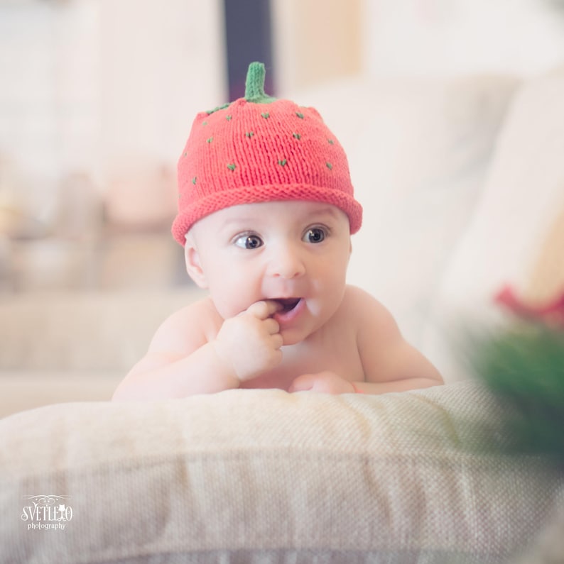Peut inclure: Un b&eacute;b&eacute; portant un bonnet en tricot rouge en forme de fraise, regardant vers la droite, avec un doigt dans la bouche.