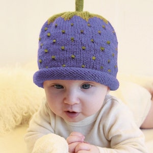 Peut inclure: Un b&eacute;b&eacute; portant un bonnet en tricot violet et vert en forme de fraise. Le bonnet a une tige verte et des graines vertes.