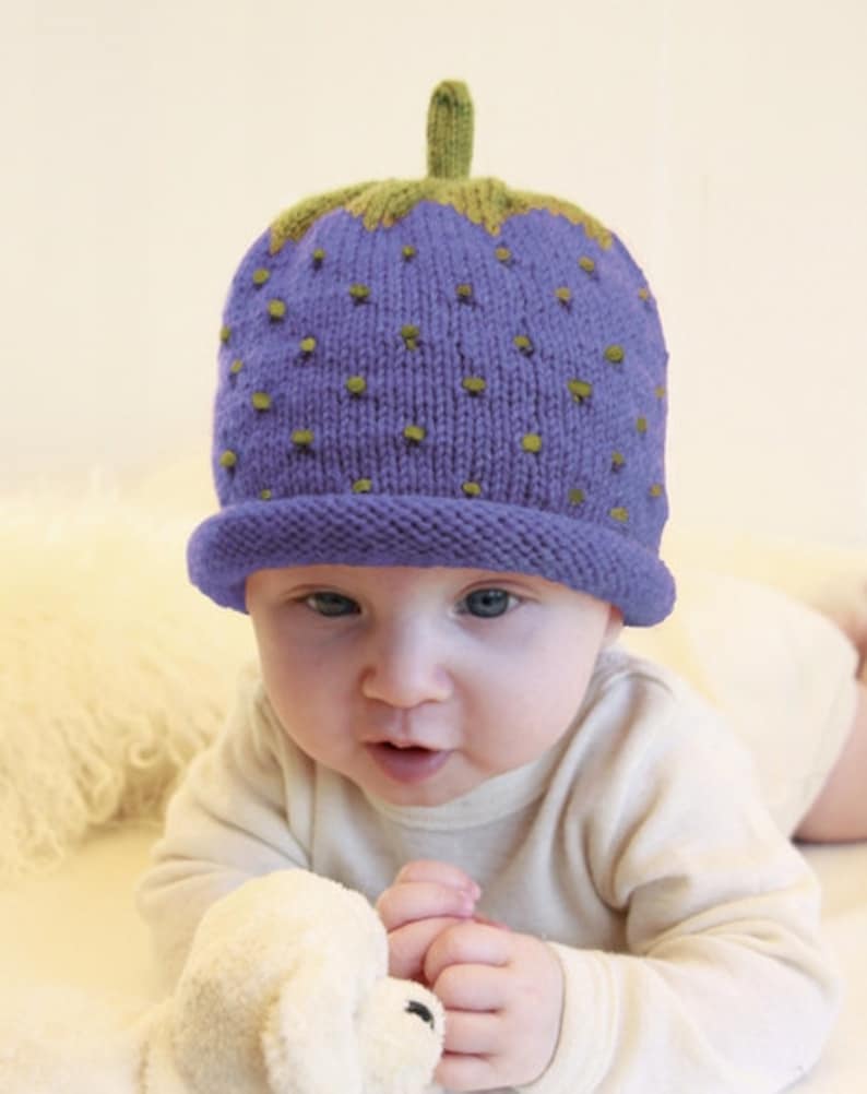 Peut inclure: Un b&eacute;b&eacute; portant un bonnet en tricot violet et vert en forme de fraise. Le bonnet a une tige verte et des graines vertes.
