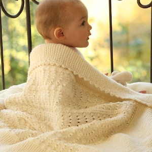 May include: A baby wearing a white knitted baby blanket with a lacy trim. The baby is sitting on a white surface and looking to the right.
