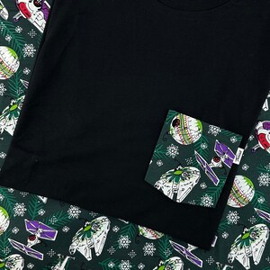 Può includere: Una maglietta nera con una tasca verde e bianca con un design natalizio con astronavi e ornamenti.