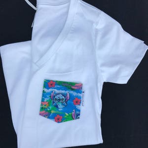 May include: White V-neck t-shirt with a blue and green pocket featuring a cartoon character with blue fur and big eyes. The pocket has a tropical print with red flowers and the text "Stitch it Up" in black.