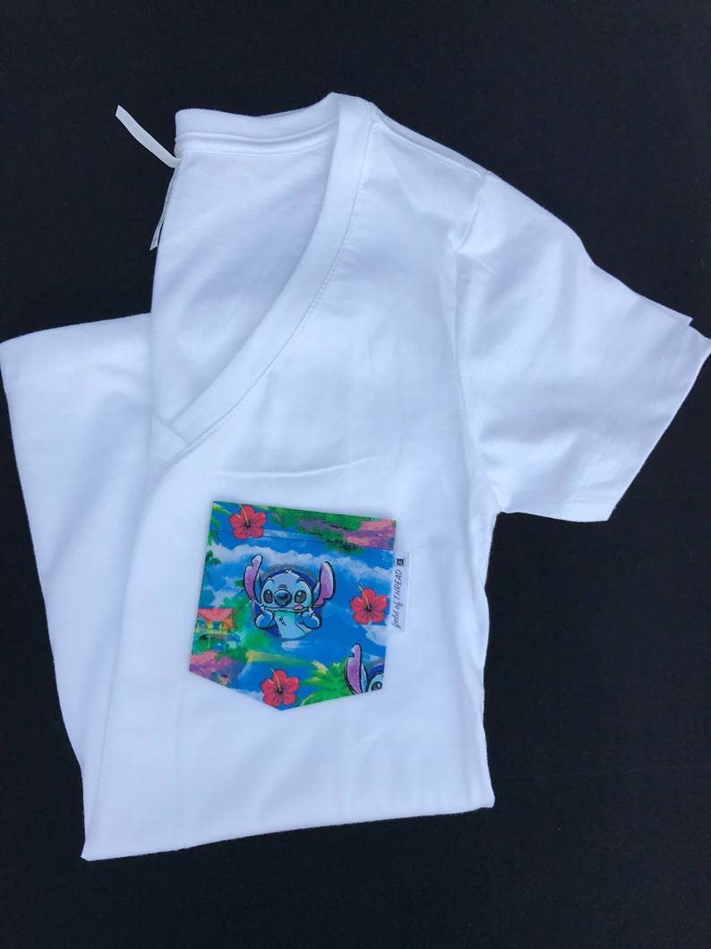 May include: White V-neck t-shirt with a blue and green pocket featuring a cartoon character with blue fur and big eyes. The pocket has a tropical print with red flowers and the text "Stitch it Up" in black.