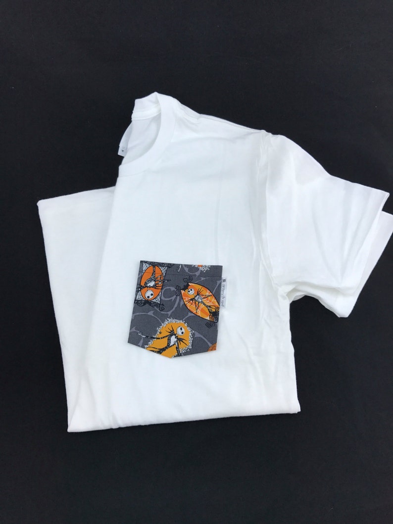 May include: White t-shirt with a pocket featuring a black and white print of Jack Skellington and Sally from The Nightmare Before Christmas. The print is in orange and grey.