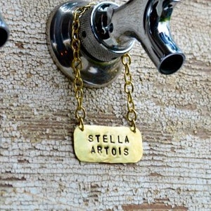 May include: A brass beer tap handle with a chain and a rectangular tag that reads "STELLA ARTOIS".