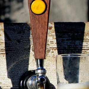 May include: A wooden beer tap handle with a yellow circular logo. The tap handle is attached to a chrome beer tap spout.