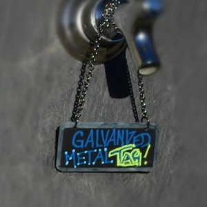 May include: A black chalkboard sign with a chain hanging from a metal tap. The sign reads "GALVANIZED METAL TAG!" in blue and yellow lettering.