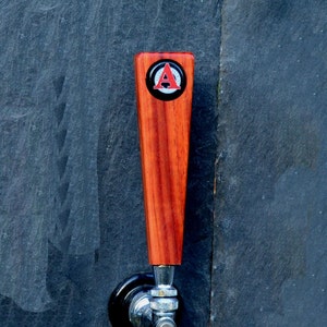 May include: A wooden beer tap handle with a red and white letter "A" on a black circle.