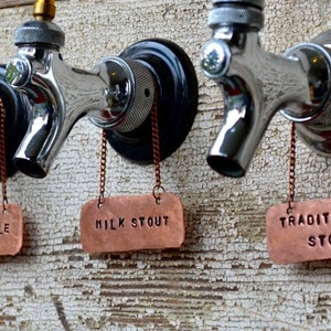 May include: Three silver beer taps with copper tags hanging from them. The tags are labeled "Pumpkin Ale", "Milk Stout", and "Traditional Stout".