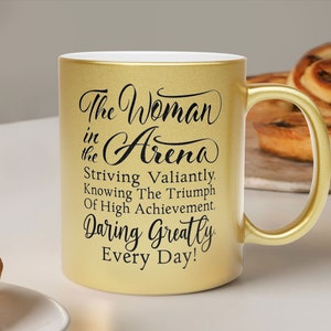 A Gold metallic mug on a white table with pastries. The mug has an abbreviated version of a Theodore Roosevelt quote, Woman in the Arena Striving Valiantly, Knowing the Triumph of High Achievement, Daring Greatly, Every Day! in black fancy font.